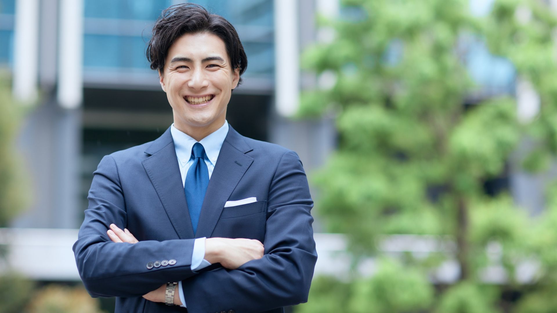 笑顔で腕を組むスーツ姿の男性がオフィスビル前に立っている写真